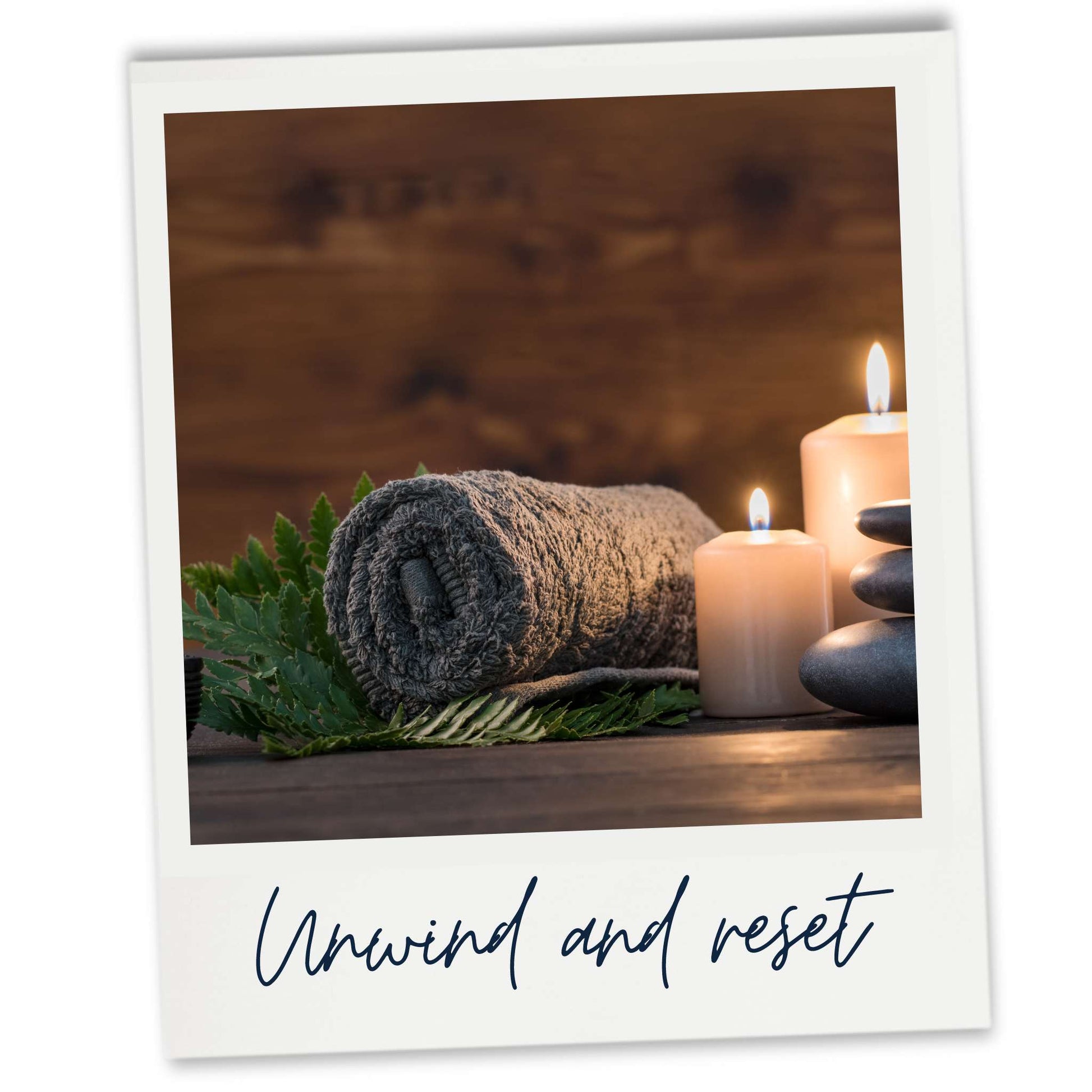 A dark grey towel rolled up on a bed of soft leaves next to lit candles and a stack of stones in a spa