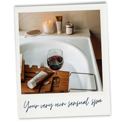 A white bathtub filled with bubbles. Surrounded by a lit candle, skincare products and a glass of red wine for an at home spa experience